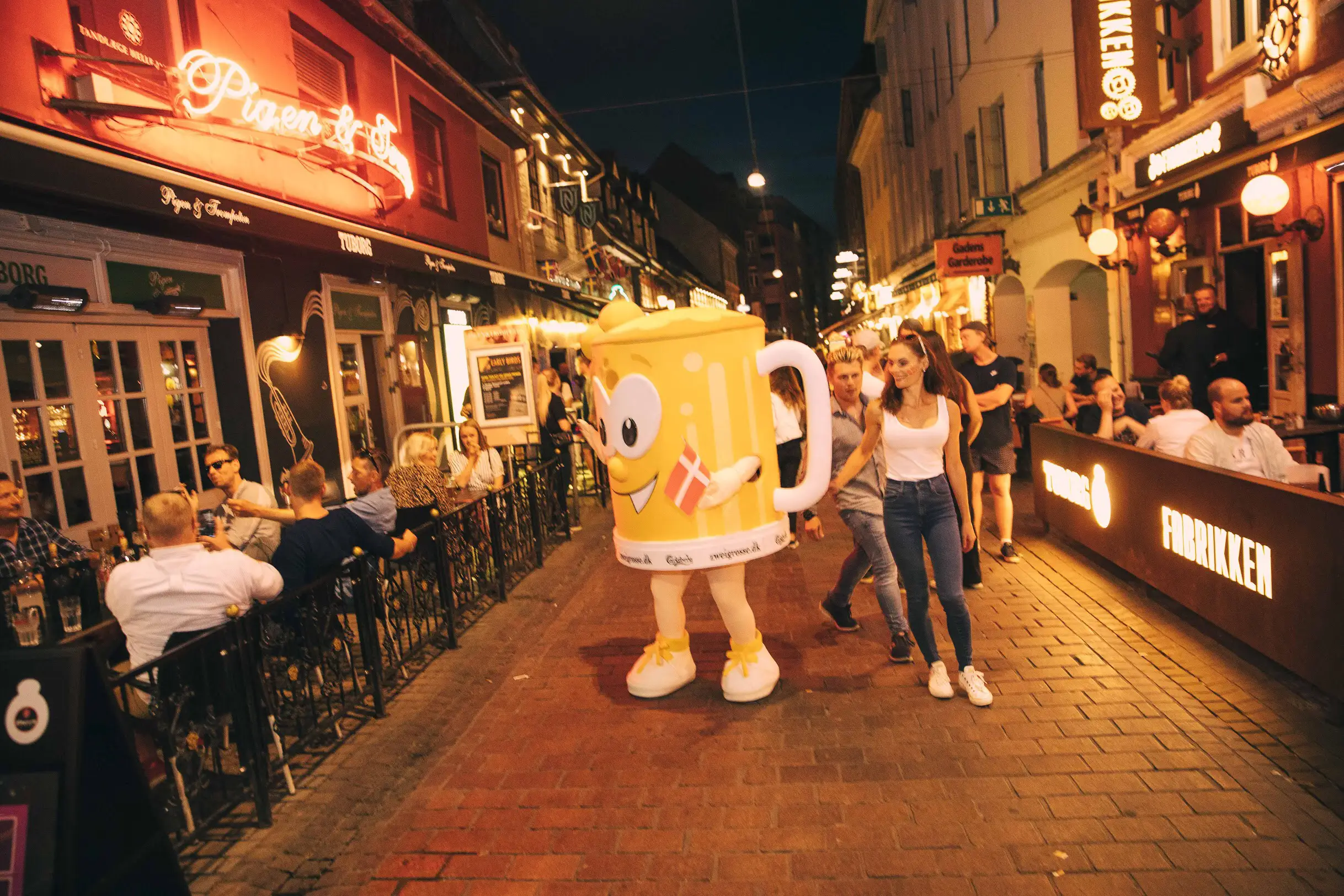 Aftenstemning i en travl udelivsgade med fortovsservering og en ølkrus-maskot